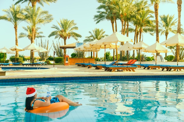 Young brunette woman in santa claus hat in a swimming circle in the pool in the Christmas holidays on the background of palm trees.