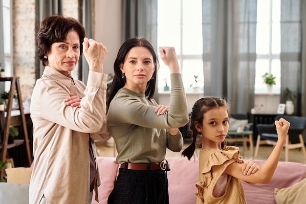 Young brunette woman, serious mature female and adorable little girl showing their strength in front of camera and looking at you