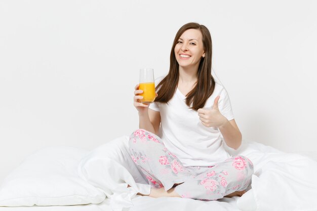 Young brunette woman sits on bed with white sheet, pillow, wrapping in blanket isolated on white wall