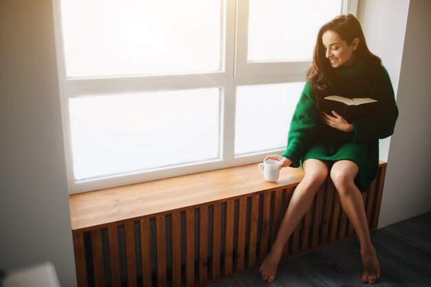 A young brunette woman sits on a windowsill and holding a book and cup of tea in her hand