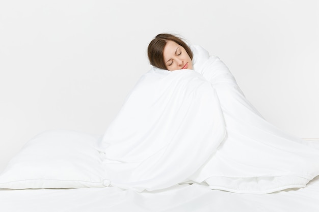 Young brunette woman sitting in bed with white sheet, pillow, wrapping in blanket on white wall