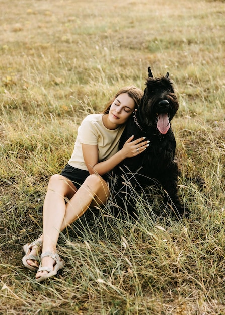 Young brunette woman sitting in a field hugging a big black dog giant schnauzer breed