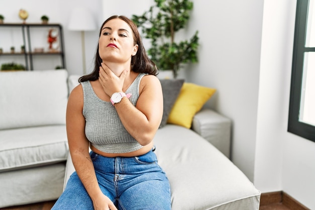 Young brunette woman sitting on the sofa at home touching painful neck, sore throat for flu, clod and infection