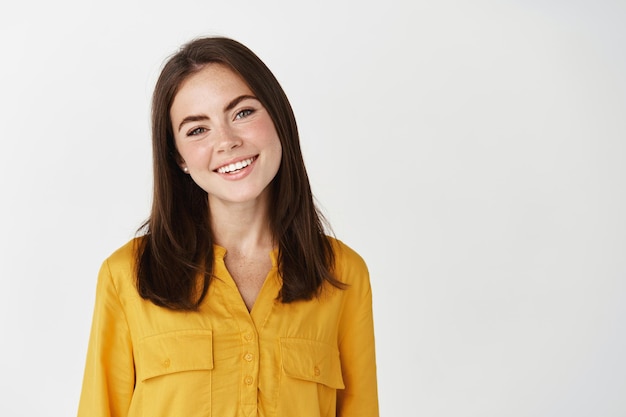 Young brunette woman smiling and looking at front, showing happy face and confidence, standing on white wall.