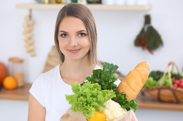 野菜や果物でいっぱいの紙袋の近くのテーブルに座って笑っている若いブルネットの女性食品購入とキッチンの寿命の概念