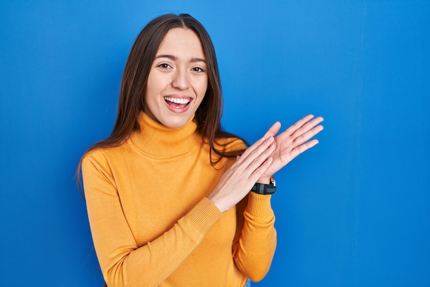 青い背景の上に立っている若いブルネットの女性の拍手と拍手幸せでうれしそうな笑みを浮かべて誇りに思って手を一緒に