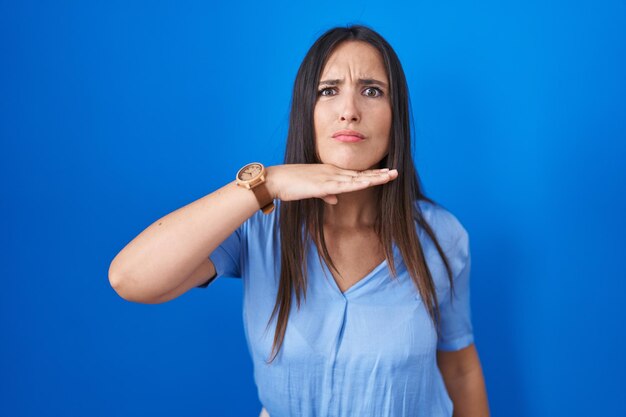 Young brunette woman standing over blue background cutting throat with hand as knife threaten aggression with furious violence