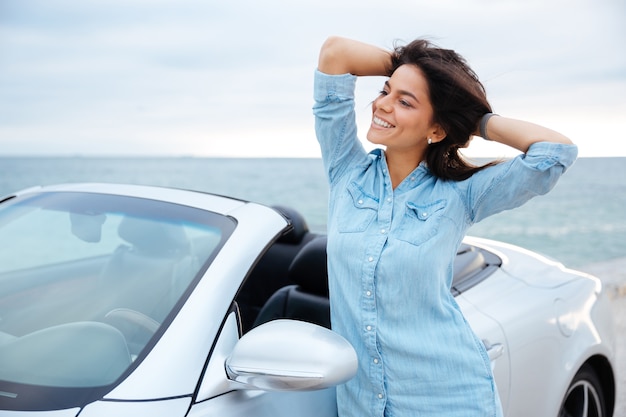 海辺で彼女の新しい車の横に立っている若いブルネットの女性