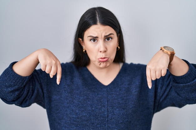 Young brunette woman standing over isolated background pointing down looking sad and upset, indicating direction with fingers, unhappy and depressed.