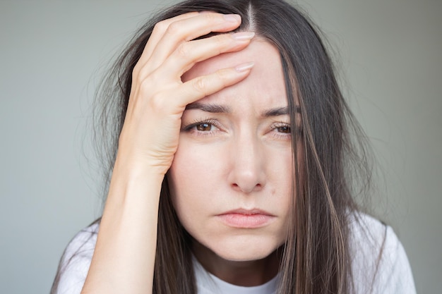 young brunette woman suffering from headache looking sadly at camera holding her head with hand