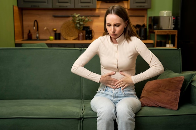 Young brunette woman suffers from abdominal pain at home sitting on a green sofa the concept of