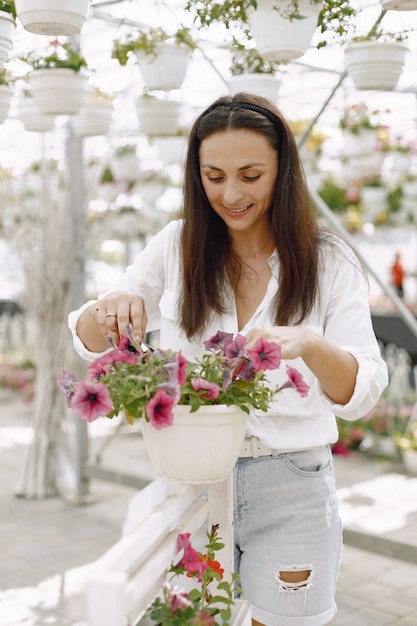 若いブルネットの女性は、庭のホースで鉢植えの植物の世話をします。白いブラウスを着ている女性