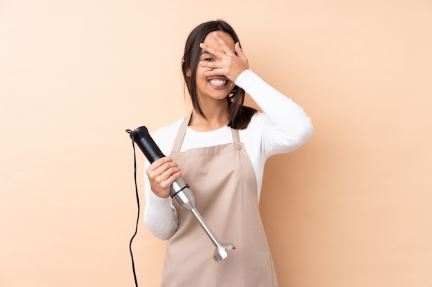 Young brunette woman using hand blender covering eyes by hands and smiling