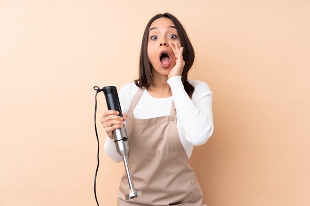 Young brunette woman using hand blender over isolated wall shouting and announcing something