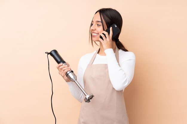 Young brunette woman using hand blender keeping a conversation with the mobile phone with someone