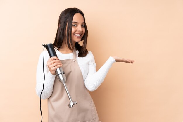 Young brunette woman using hand blender presenting an idea while looking smiling towards