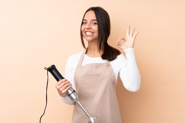 Young brunette woman using hand blender showing ok sign with fingers