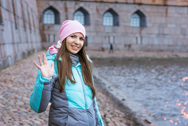 Young brunette woman in warm clothes and pink hat stands by river and smiles bright colored outerwea...