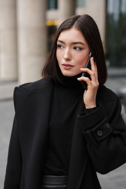 young brunette woman wearing autumn coat standing in a street, using mobile phone