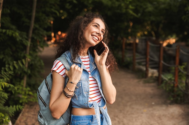 молодая брюнетка женщина в рюкзаке, улыбается во время прогулки по зеленому парку и разговаривает по смартфону