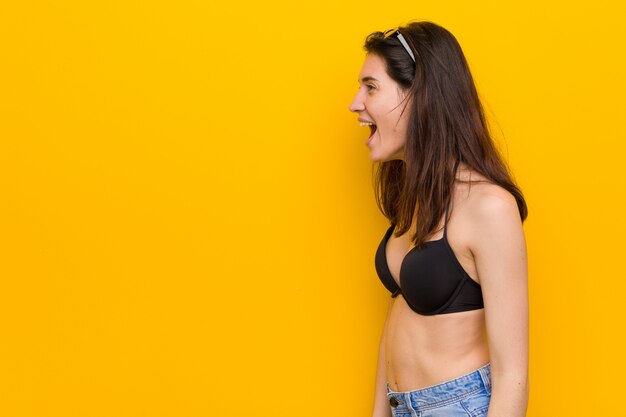 Young brunette woman wearing a bikini against yellow background shouting