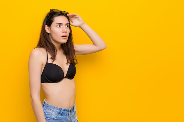 Young brunette woman wearing a bikini against yellow wall looking far away keeping hand on forehead.