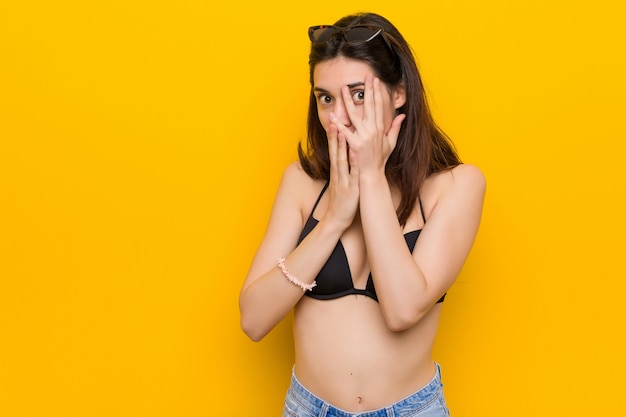 Young brunette woman wearing a bikini on yellow blink through fingers frightened and nervous.