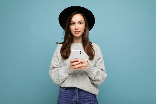 Young brunette woman wearing black hat and grey sweater holding smartphone looking at camera