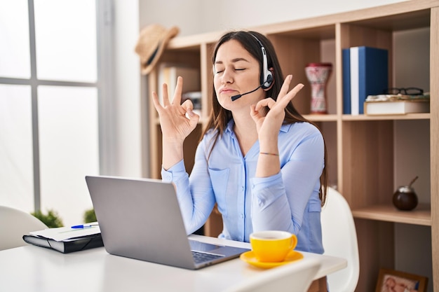 Young brunette woman wearing call center agent headset relax and smiling with eyes closed doing meditation gesture with fingers. yoga concept.