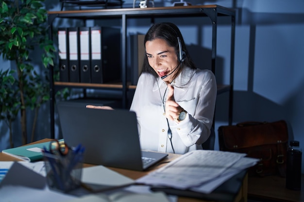 Young brunette woman wearing call center agent headset working late at night looking at the camera smiling with open arms for hug cheerful expression embracing happiness