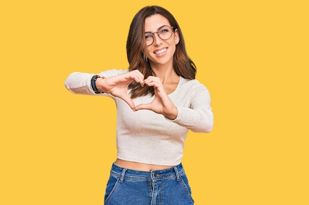 Foto giovane donna castana che indossa abiti casual e occhiali sorridenti innamorati che fanno la forma del simbolo del cuore con il concetto romantico delle mani