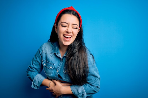 Young brunette woman wearing casual denim shirt over blue isolated background smiling and laughing hard out loud because funny crazy joke with hands on body