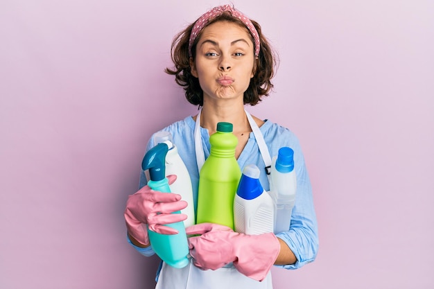 Young brunette woman wearing cleaner apron holding cleaning products puffing cheeks with funny face mouth inflated with air catching air