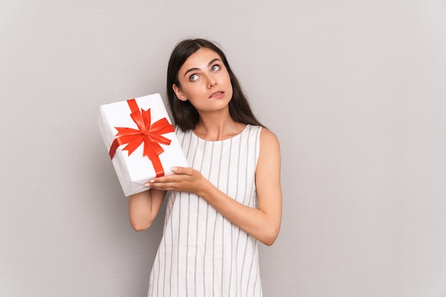  young brunette woman wearing dress thinking while holding present box isolated over gray wall