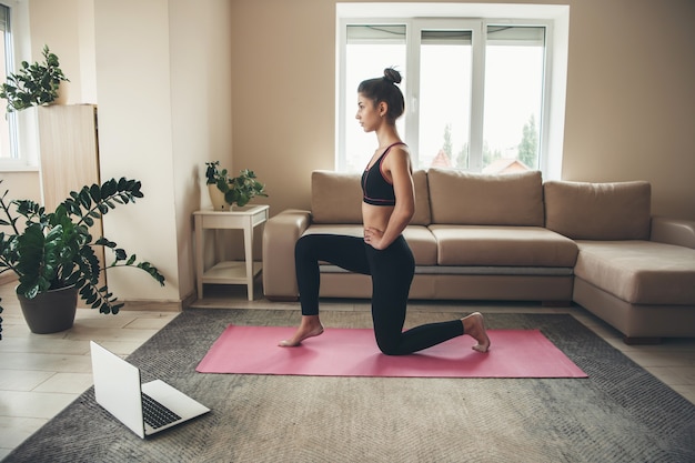 Young brunette woman wearing sportswear is doing yoga on a pink carpet using a laptop