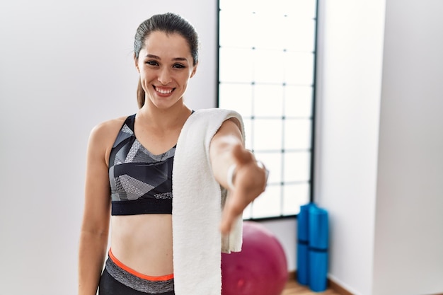 Young brunette woman wearing sportswear and towel at the gym smiling friendly offering handshake as greeting and welcoming successful business