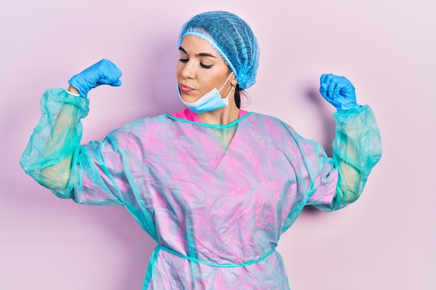 Young brunette woman wearing surgeon uniform and medical mask showing arms muscles smiling proud fitness concept