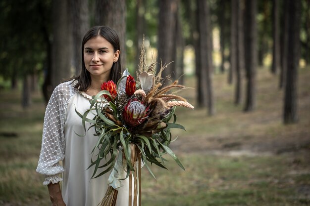 Young brunette woman in a white dress with a bouquet of flowers in the forest on a blurred background, copy space.