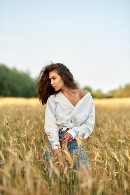 黄金の麦畑を背景に白いシャツとブルージーンズの若いブルネットの女性