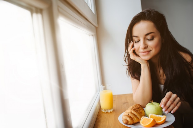 Молодая брюнетка женщина в белом белье завтрака на деревянный подоконник. Молодая женская модель ест.