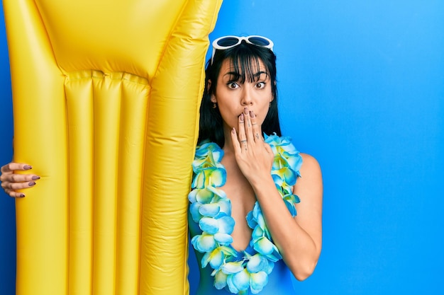 Young brunette woman with bangs wearing swimsuit and holding summer float covering mouth with hand shocked and afraid for mistake surprised expression