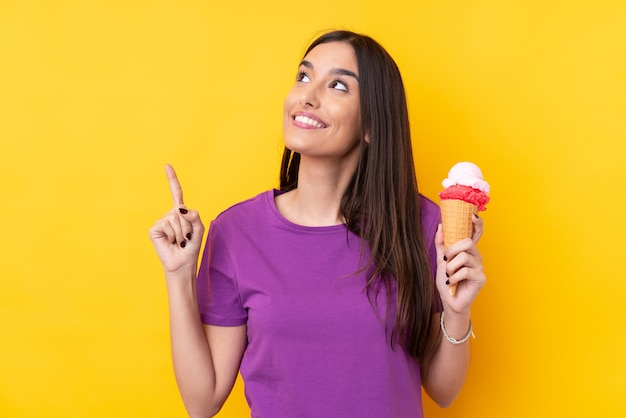 Young brunette woman with a cornet ice cream over isolated yellow wall intending to realizes the solution while lifting a finger up