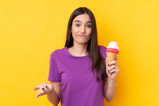 Young brunette woman with a cornet ice cream over isolated yellow wall making doubts gesture while lifting the shoulders