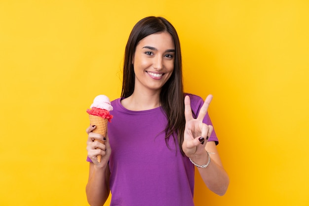 笑顔と勝利のサインを示す分離の黄色の壁の上のコルネットアイスクリームの若いブルネットの女性