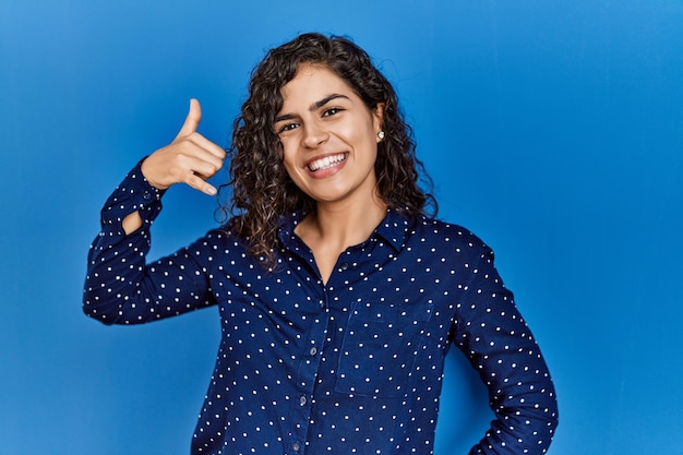 Young brunette woman with curly hair wearing casual clothes over blue background smiling doing phone gesture with hand and fingers like talking on the telephone communicating concepts