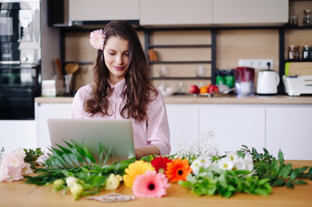 Foto giovane donna bruna con lunghi capelli ondulati che lavora al computer portatile con fiori sulla scrivania con cucina sullo sfondo fiorista di talento che sviluppa vendite online e si prepara per l'officina