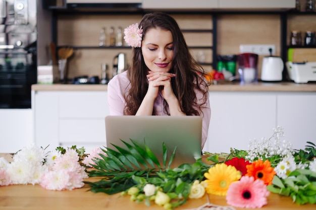 Giovane donna bruna con lunghi capelli ondulati che lavora al computer portatile con fiori sulla scrivania con cucina sullo sfondo fiorista di talento che sviluppa vendite online e si prepara per l'officina