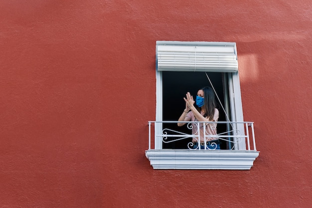 Foto giovane donna castana con la maschera che applaude dalla finestra della casa