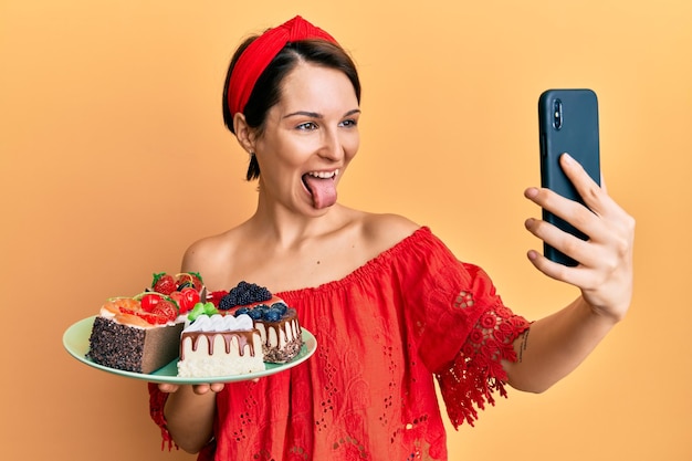 Young brunette woman with short hair holding sweets cakes taking a selfie picture sticking tongue out happy with funny expression