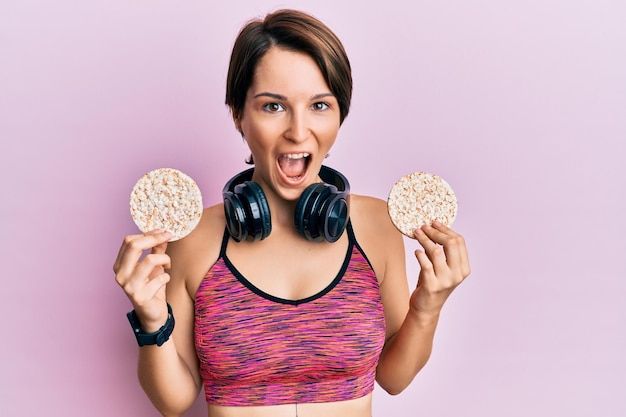 Young brunette woman with short hair wearing sportswear holding rice cakes celebrating crazy and amazed for success with open eyes screaming excited